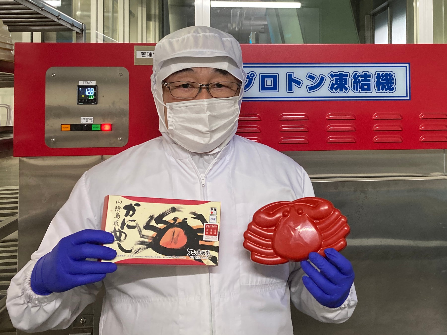 *プロトン凍結機の前で、冷凍グルメ駅弁「山陰鳥取かにめし」を持つ、アベ鳥取堂の阿部社長