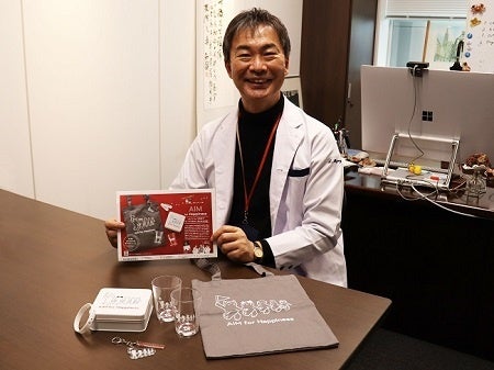 猫部から販売中のAIM医学研究支援基金付きチャリティーグッズと宮崎徹氏(2023年3月一般社団法人AIM医学研究所にて)