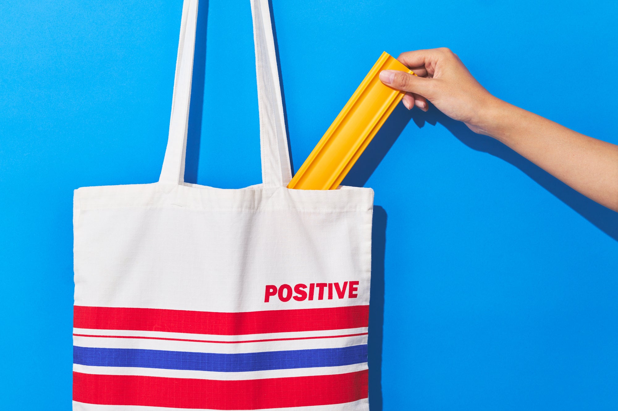 画像の説明：ココテープのビジュアル。水色の背景に、赤と青のストライプのトートバックが。右側から手が伸びており、バックから黄色いココテープを取り出そうとしている。