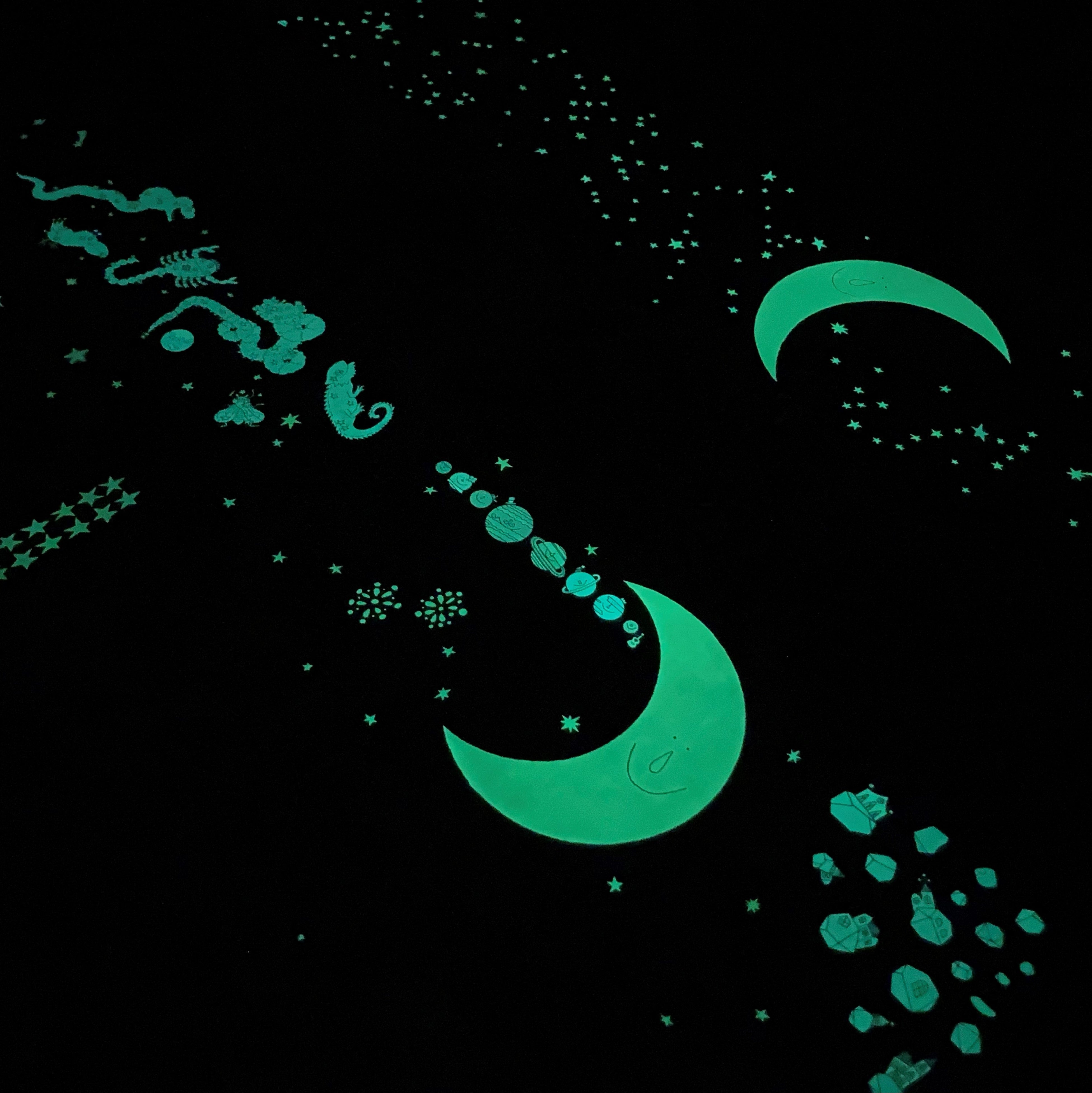 ▲電気を消すと、こんな風に月と星部分が、青白く幻想的に光ります！　『おつきさまのおさんぽ 新版』蓄光ポスター（右奥）、『おつきさまのともだち 新版』蓄光ポスター（左手前）