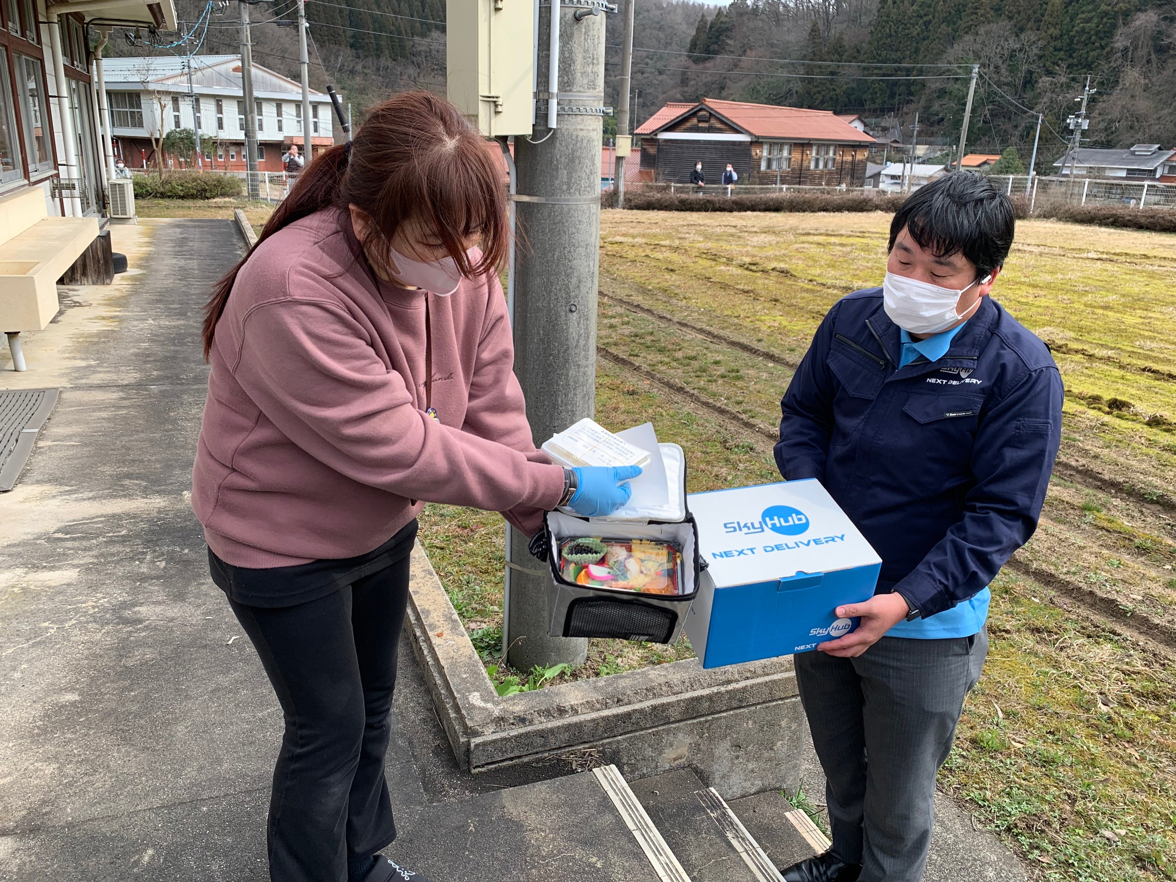 ドローン配送された配食サービスのお弁当を受取る介護事務所のスタッフ （入間介護事務所前）