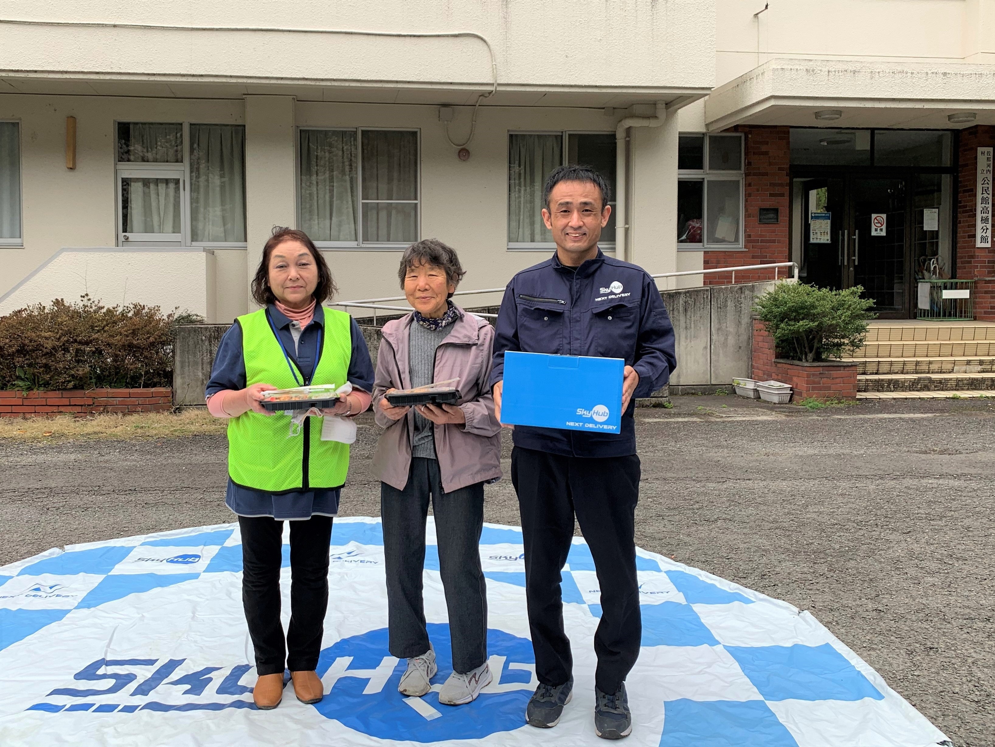 日本発物流専用ドローン“AirTruck”で配送された２人分のお弁当とお茶を受け取った住民と記念撮影