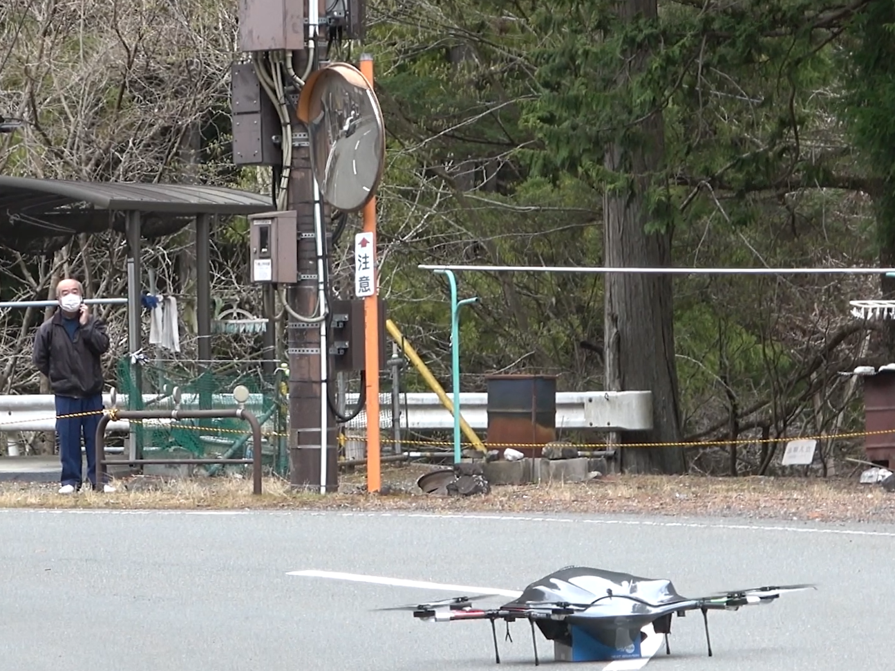 中津川区長が、東京の遠隔運用者と離陸地点スタッフの3者間通話で着陸地点の状況を確認する中、ドローンが着陸 （崩落地点を超えた先の着陸地点）