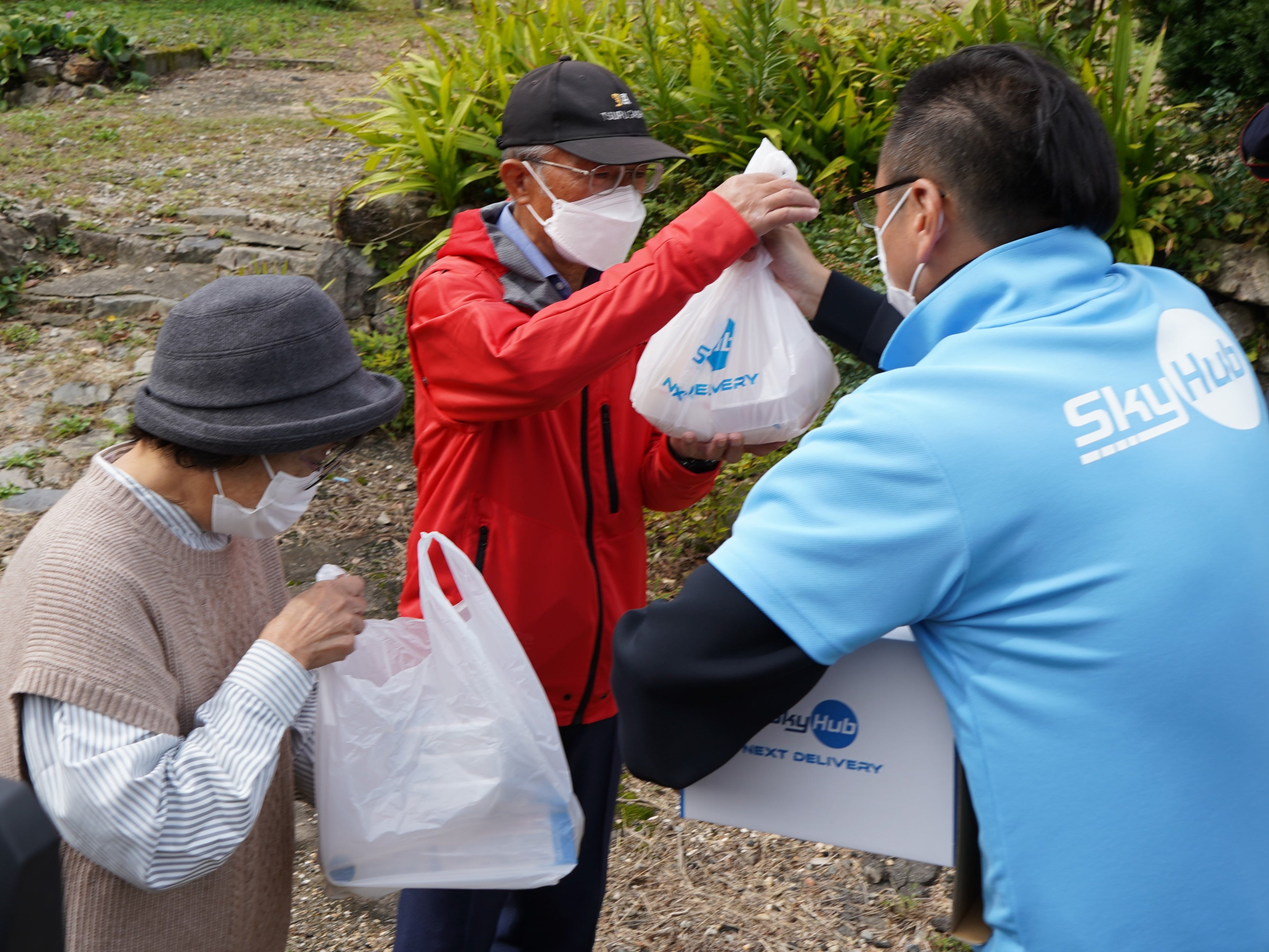SkyHub®Storeサービスでドローン配送された商品をスタッフから受取る杉本杉箸区長と早瀬さん（杉箸ドローンスタンド®脇）