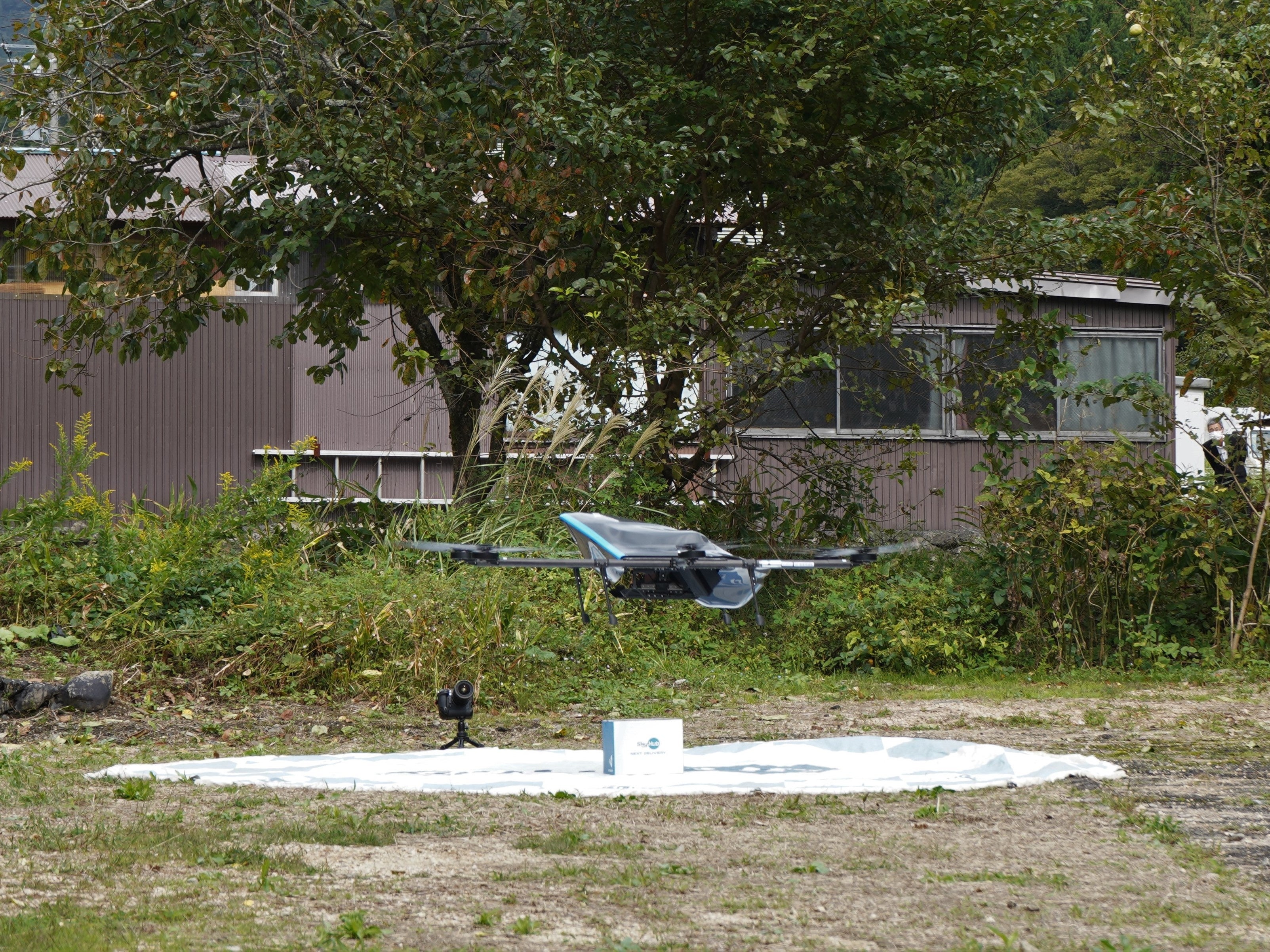 SkyHub®Storeサービスでドローン配送された商品の入った箱を置いて「ドローンデポ®愛発」に戻るドローン（杉箸ドローンスタンド®）