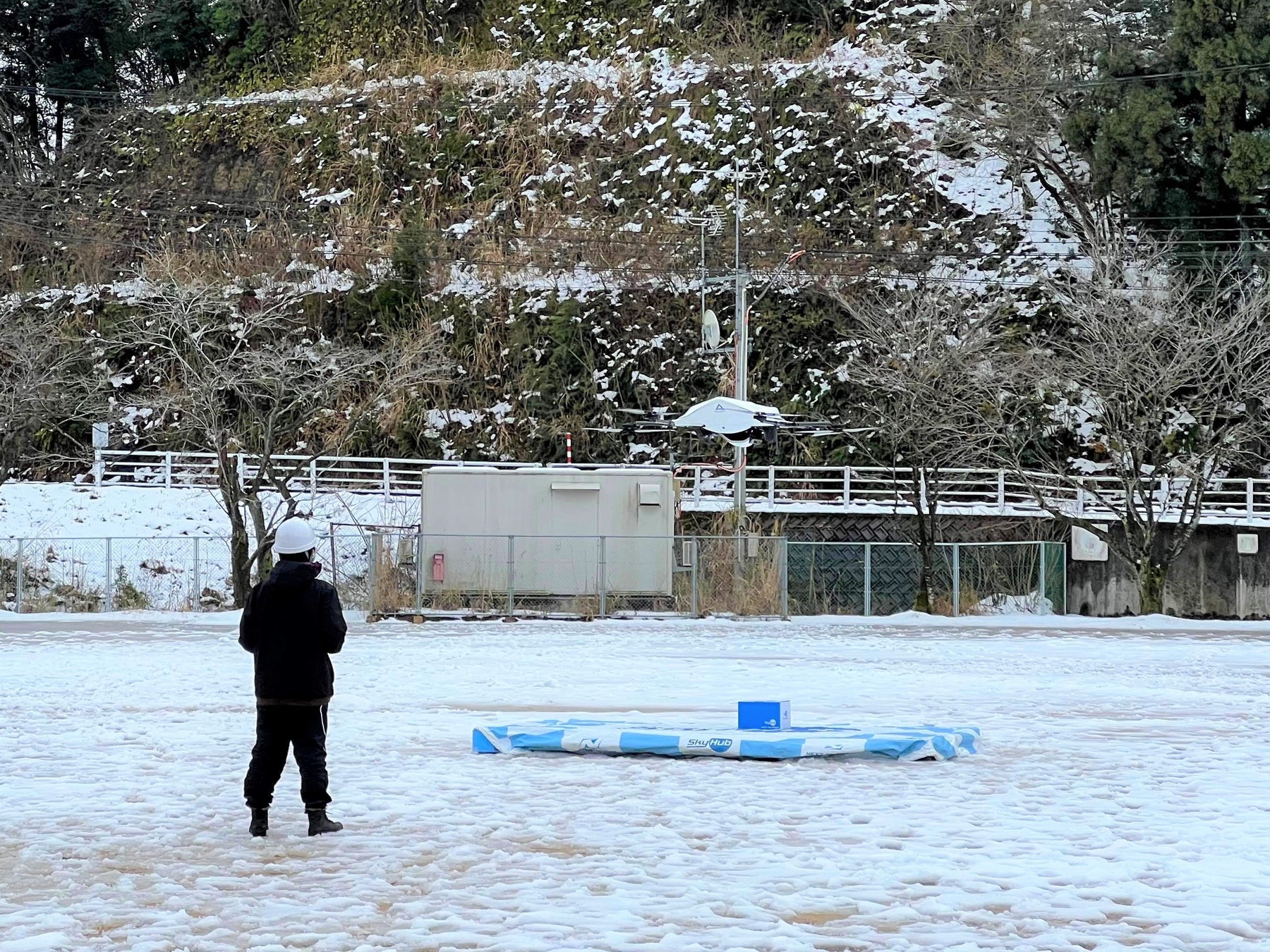 雪景色の中、児童生徒への荷物を届けて飛び立つドローン （松東みどり学園グラウンド）