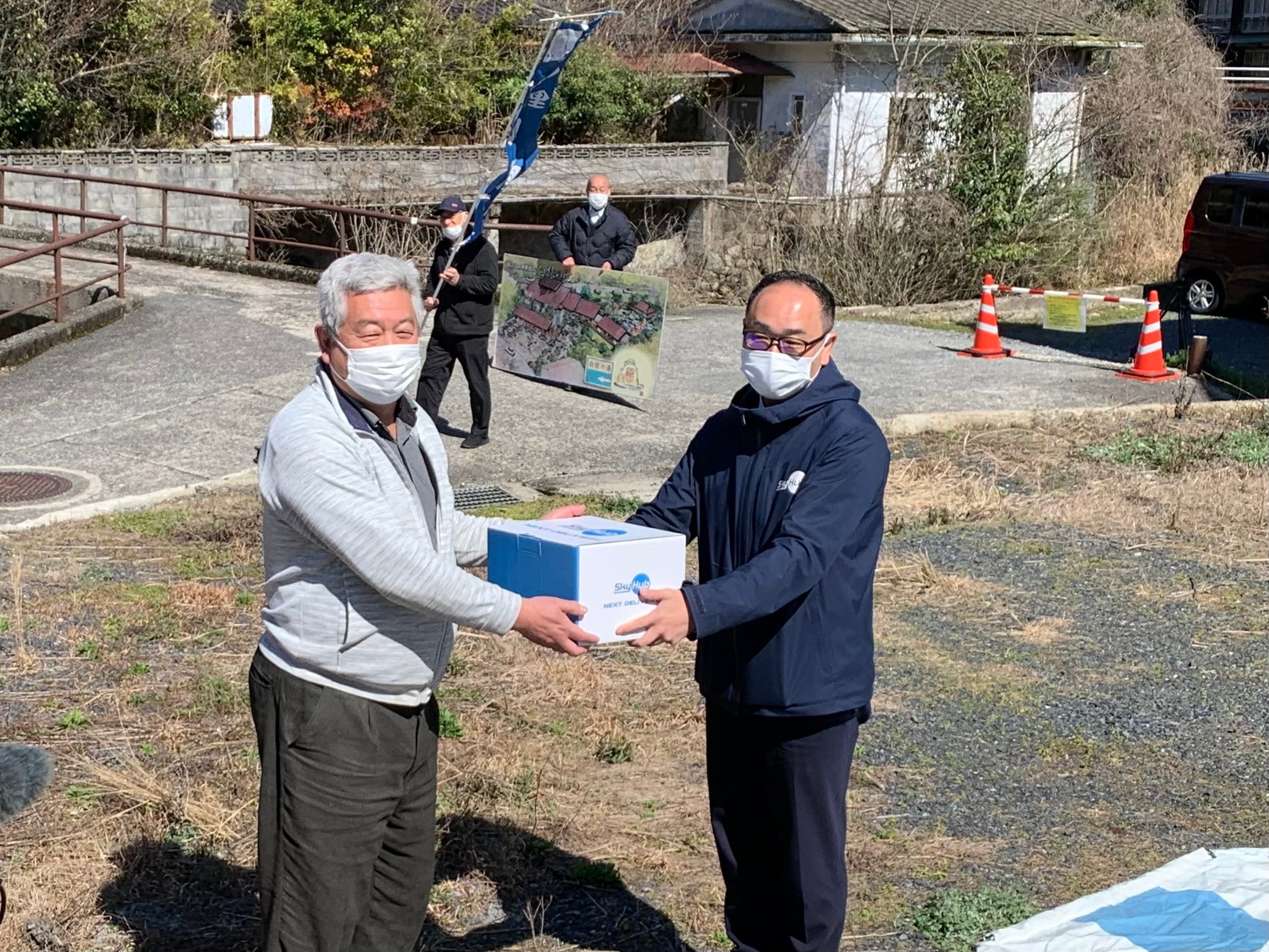 ドローン配送された商品を受取る ゲストハウスのオーナー（写真左） （俵山地区）