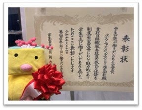 表彰状とMAKE HAPPY プロジェクトのマスコットキャラクター「はっぴーさん」（左）