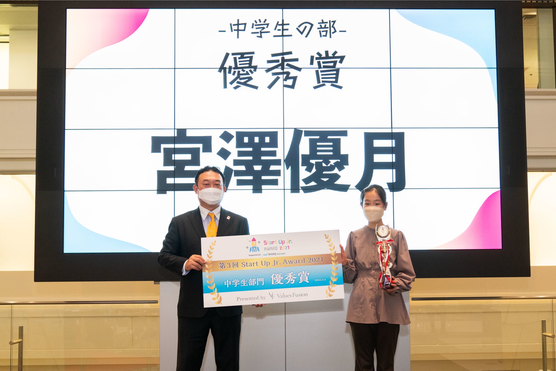 審査員の(公社)2025年日本国際博覧会協会 広報戦略局長 堺井 啓公氏よりトロフィーとパネルを授与される宮澤さん