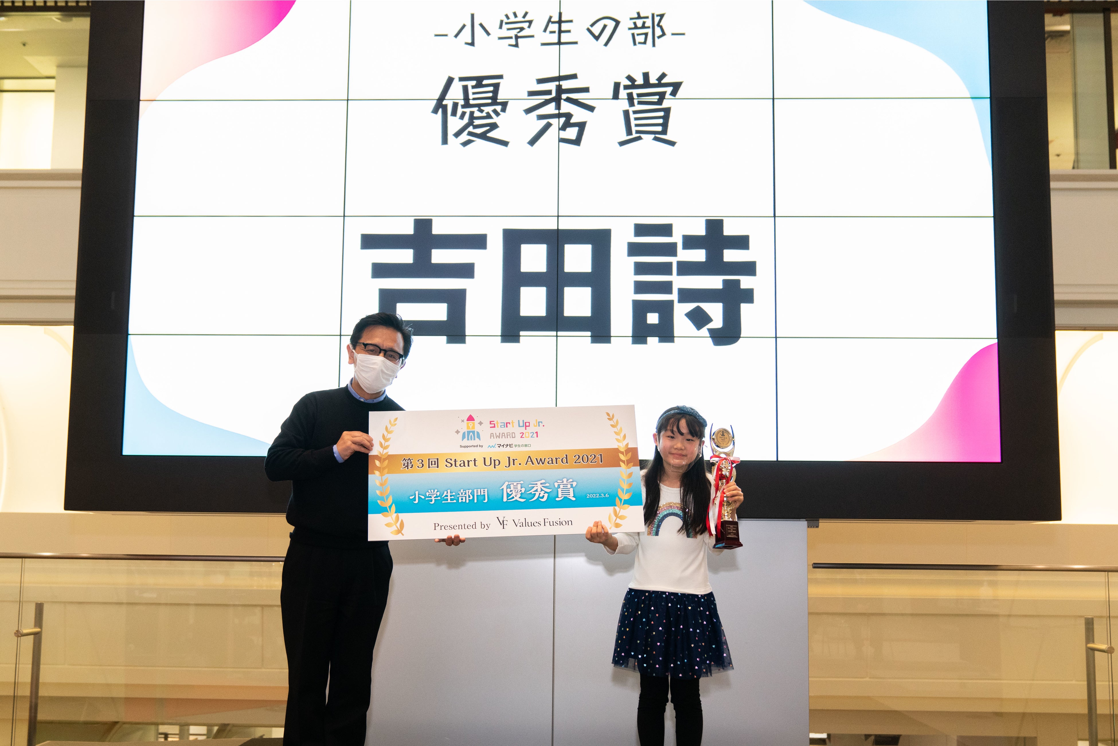 審査員の（株）ウェルカム代表取締役&nbsp;&nbsp;横川 正紀氏 よりトロフィーとパネルを授与される吉田さん