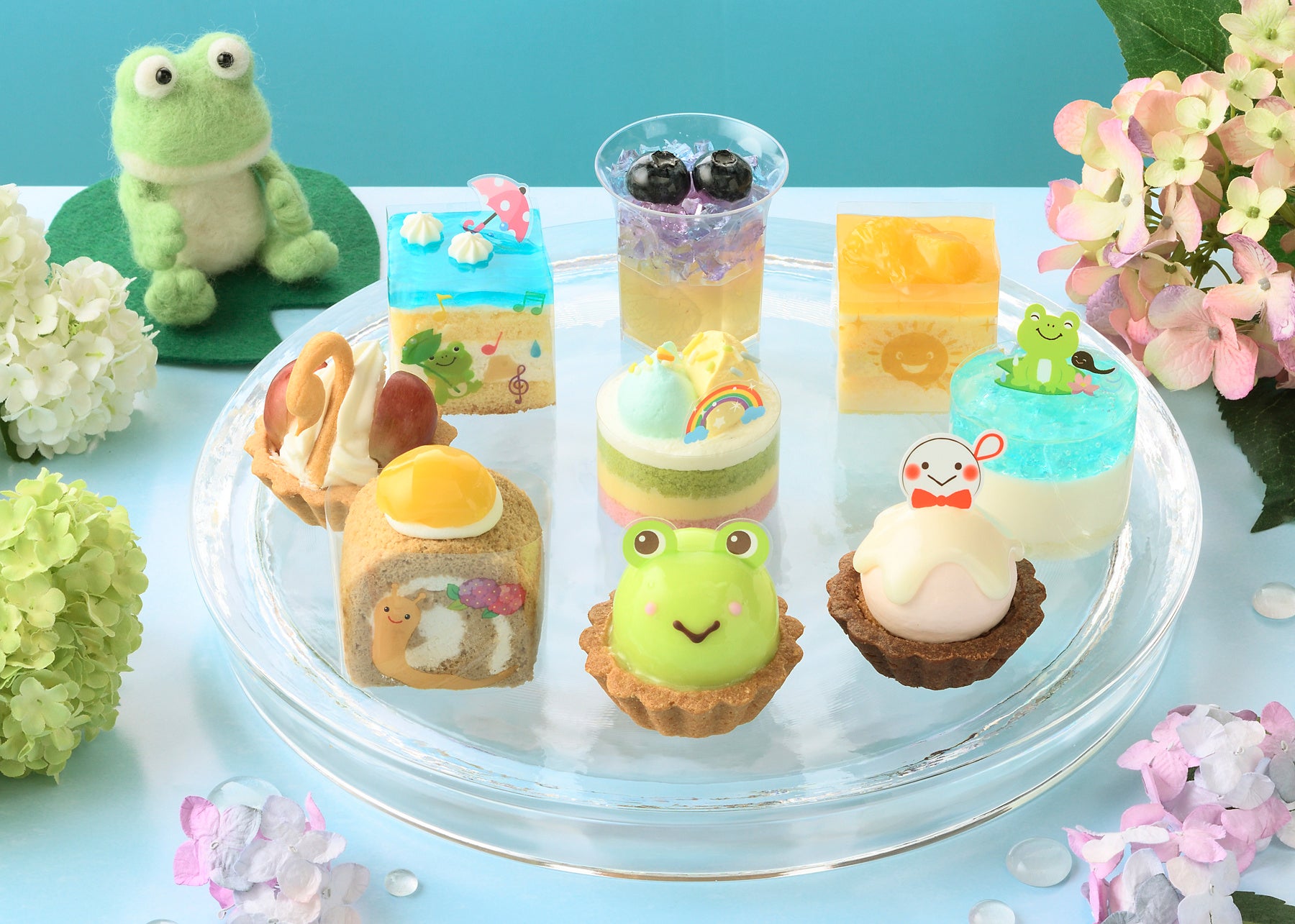 透明なガラス皿に並べたプチケーキ9個のイメージ。写真左奥のフェルトのカエルは、今回も当商品の企画開発担当者が作成。ストーリーや挿絵、キャラクターなどの制作もすべて当社社員が手掛けています。