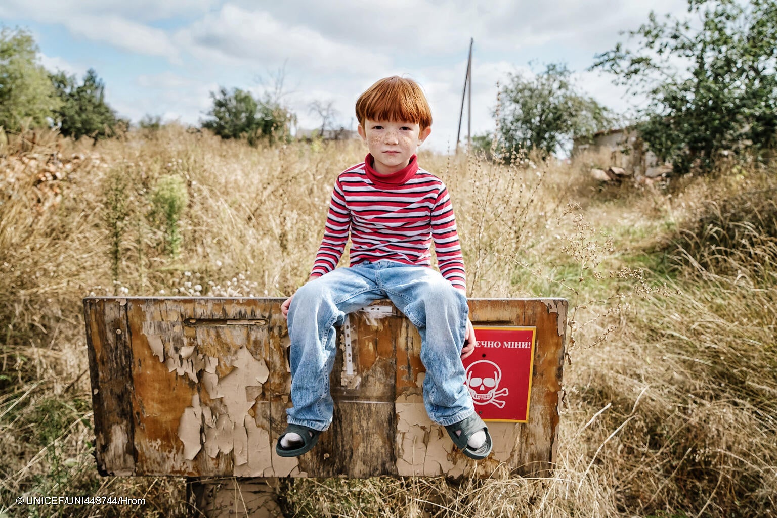 「地雷の危険」と書かれた看板の上に座る6歳のレオニードさん。戦闘が激化しても家族はハルキウ州の自宅に留まり続けた。地元の学校は破壊されてしまったため、レオニードさんはオンラインで授業を受けている。(ウクライナ、2023年9月撮影) © UNICEF_UNI