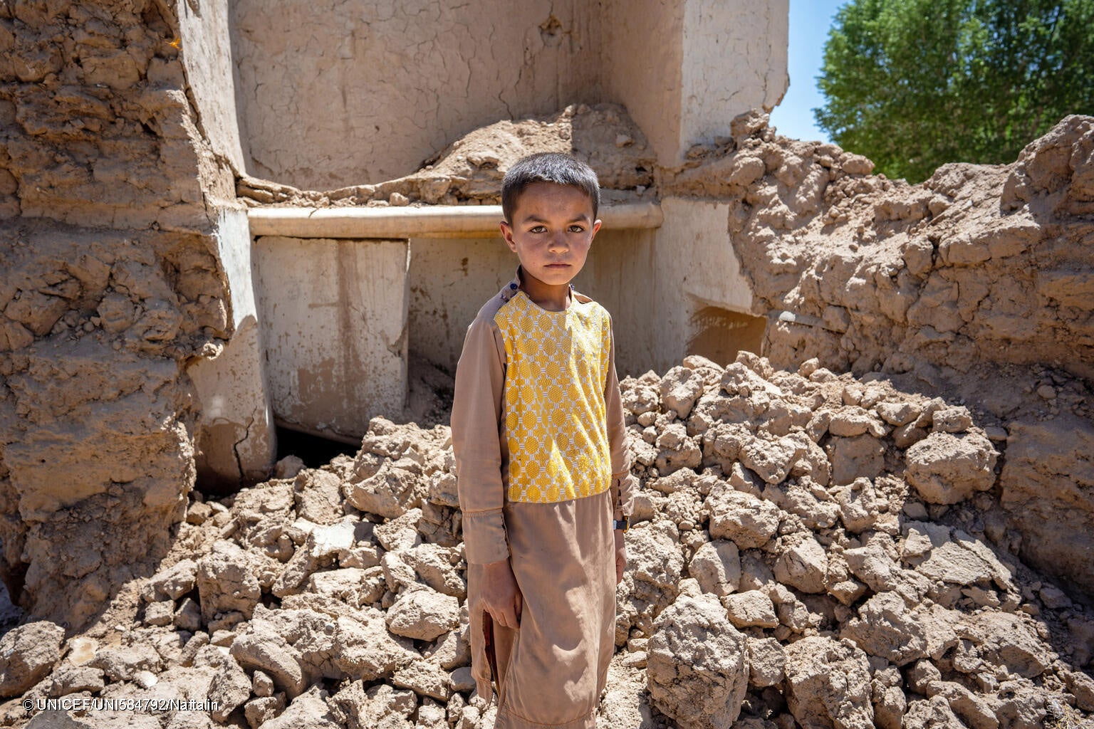 西部ゴール州で、洪水により破壊された自宅前に立つ9歳のファルザンさん。持ち物をすべて失ったため、いとこの服を借りて着ている。(アフガニスタン、2024年5月28日撮影) © UNICEF_UNI584792_Naftalin