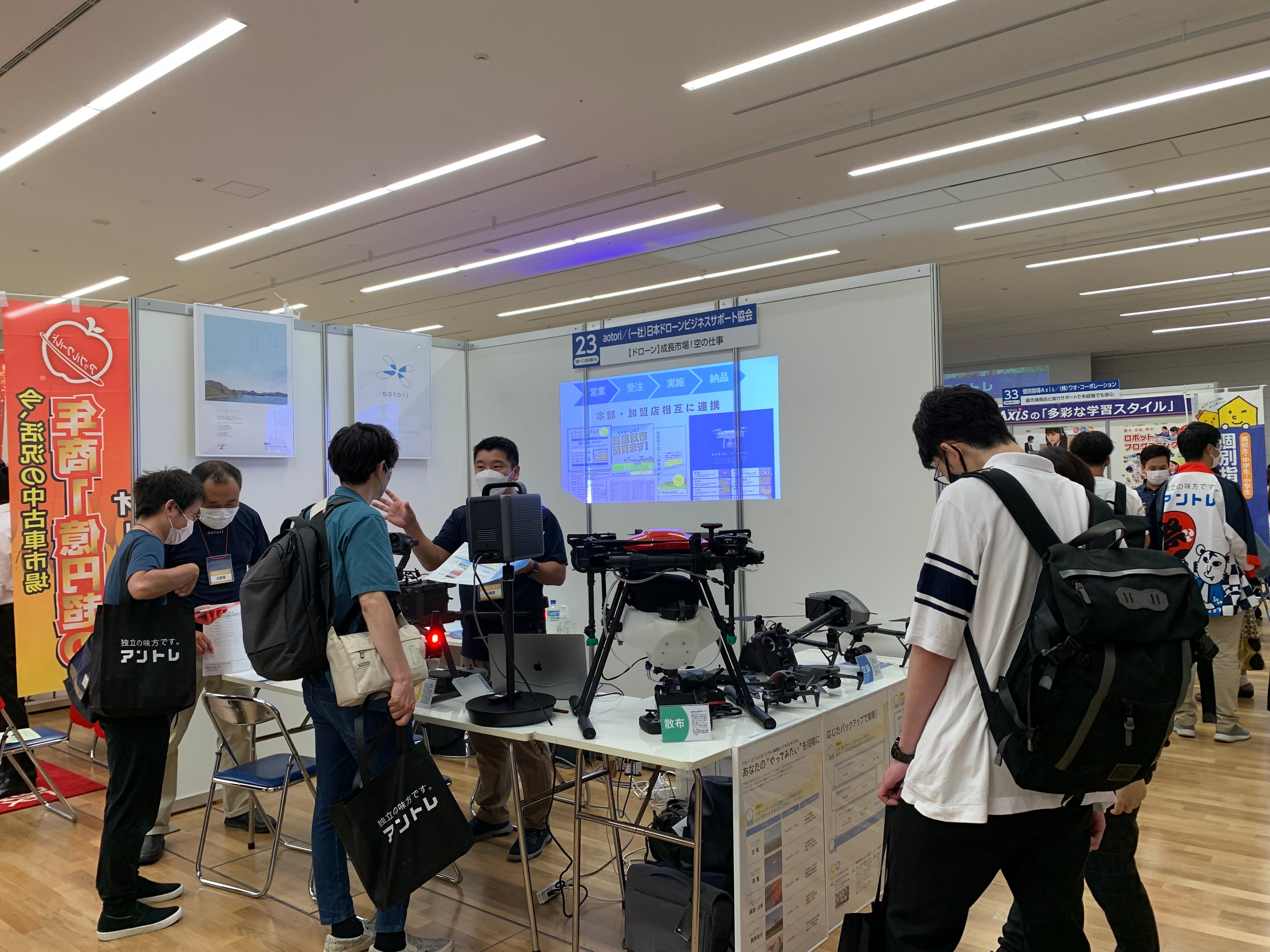 ドローンビジネスについて紹介する出展企業 （一般社団法人日本ドローンビジネスサポート協会）と説明を聞く来場者