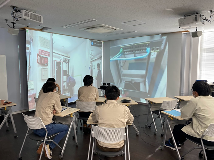 左側スクリーンで作業空間を共有し、かつ、スマートグラスで作業者の視点を右側スクリーンにリアルタイムに配信することで、実機ランプの点灯状況確認を疑似体験している様子