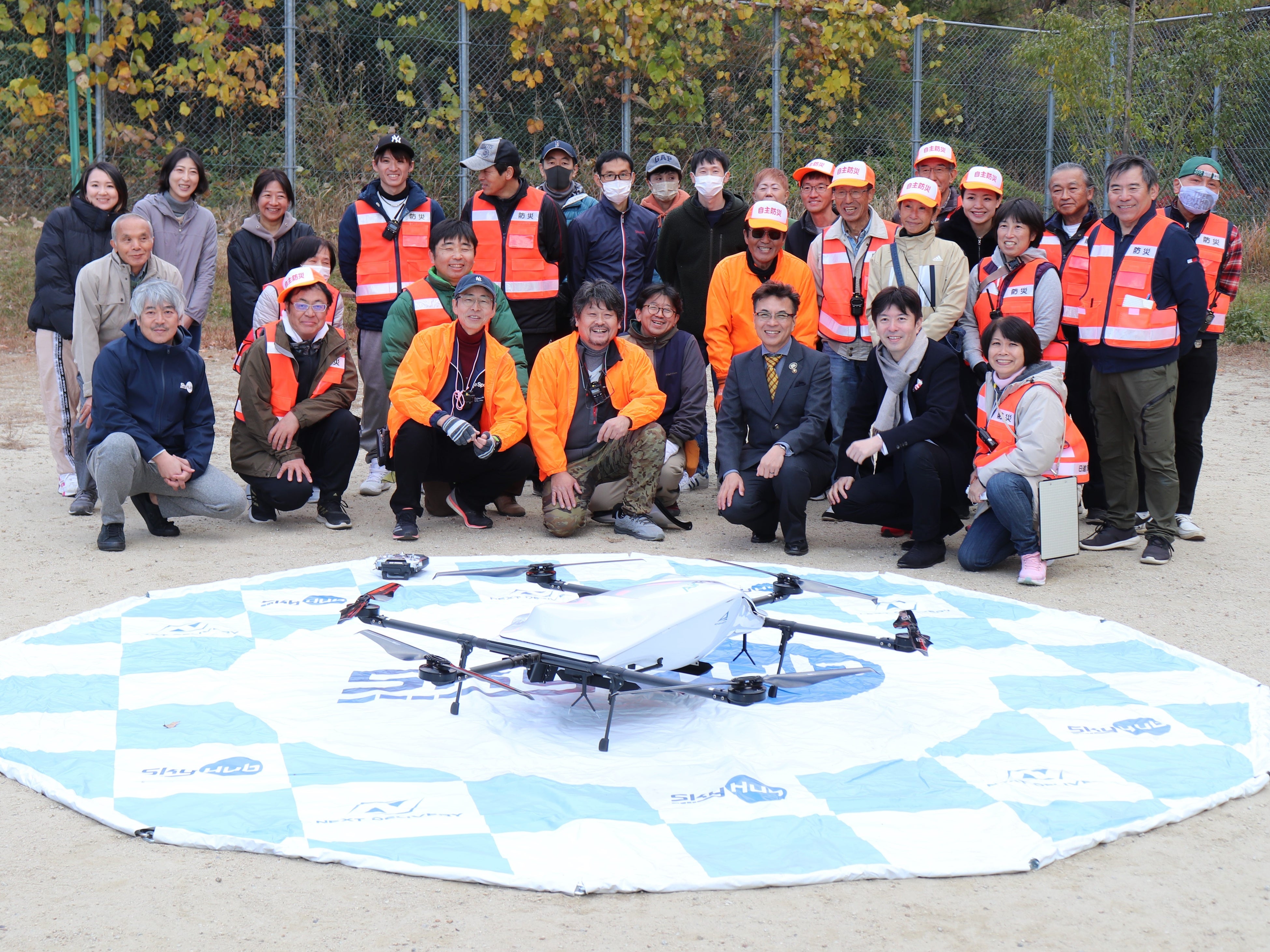 配送を終えたドローンを前に北山ふれあい広場に集まった日進ニュータウン自主防災会関係者と住民の皆さんと記念撮影 （北山ふれあい広場）